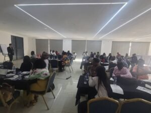 A presenter in a suit speaks at the front of a bright conference room to a large audience seated at multiple tables.