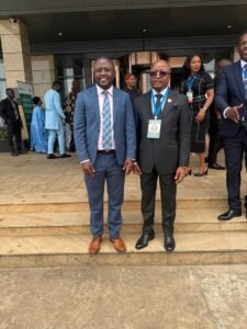 Two men in suits pose on building steps at a formal event, with others in the background wearing badges and lanyards.