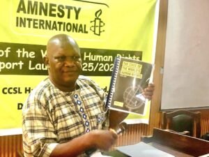 Man in a checked shirt holds a spiral-bound book titled 'The State of the World's Human Rights' in front of an Amnesty International banner at a rights event.