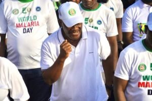 Smiling man in a white polo and cap taking part in a World Malaria Day event, with others in matching shirts in the background.