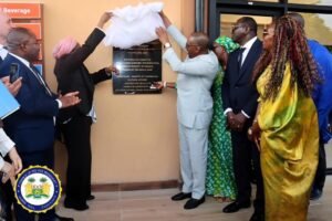Officials unveil a plaque mounted on a beige wall during a formal ceremony, with diverse attendees in suits and dresses down the line of people present at the event.