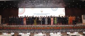 Group of formally dressed delegates standing on a stage for a summit photo, with a banner reading 'India Africa Forum Summit-IV' behind them and flower decorations in front.