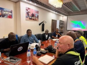 Group of men seated around a wooden conference table in a meeting room, with laptops, notebooks, and water bottles; a colorful projector image lights the screen in the background.