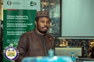 Man in traditional attire speaking at a podium during a ECOWAS Judicial Council statutory meeting, microphone in front of him.