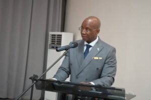 Bald man in a gray suit speaks at a podium with a microphone during a formal event, wearing glasses and a blue tie.