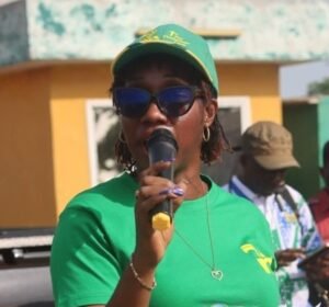 Woman in a green shirt and cap speaks into a handheld microphone at an outdoor community event, wearing sunglasses and hoop earrings.
