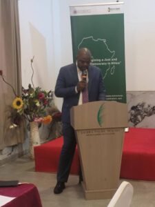 Man in a blue suit giving a speech at a wooden podium in a conference room, holding a microphone; a green banner with an outline of Africa and event text stands behind him, with floral arrangements on the left and red carpeted stage.