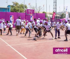 Group of youths playing basketball on an outdoor court, wearing white shirts; purple branding banners and tents surround the court, with an Africell logo visible in the corner.