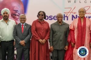 Five adults in formal attire posing for a photo at a welcome-event backdrop, with a circular presidency seal in the corner.