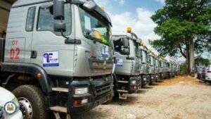 A row of gray heavy‑duty trucks parked on a dirt lot under a blue sky with trees in the background.