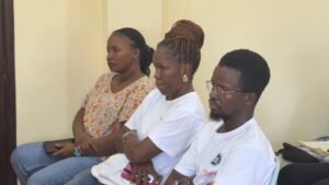 Three people sit side by side in a pale hallway, listening intently to something off-camera.