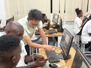 Instructor guides a student at a computer in a busy classroom tech lab.