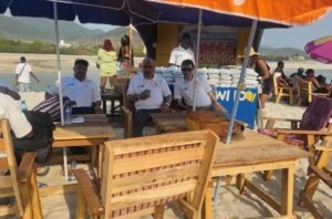 Four people in white shirts sit at wooden tables under blue and orange beach umbrellas on a sunny sandy beach during an event, with a banner and other attendees in the background.