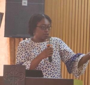 Woman wearing a blue and white patterned dress speaks into a handheld microphone at a podium.