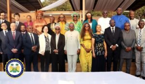 Group of diverse officials in suits and traditional attire posing for a formal photo at an outdoor event, with a government seal visible on the table front left.