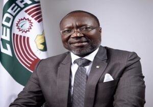 Professional man in a dark suit with a white pocket square, seated with a colorful flag in the background.