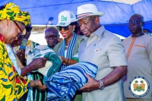 An official in a light gray suit hands a folded blue striped blanket to an elderly woman in a bright yellow dress under a blue canopy, with onlookers nearby.