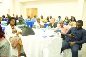 Diverse group of attendees seated around round tables in a conference room, with water bottles, notebooks, and phones visible.