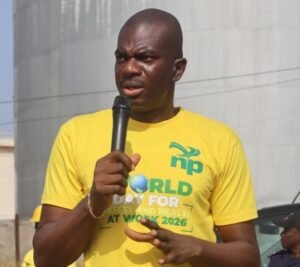 Man in a yellow event T-shirt speaks into a handheld microphone outdoors at a public gathering.