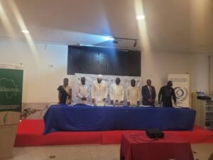 Seven panelists stand behind a long blue-draped table on a red-carpeted stage, with banners on either side during a formal conference.