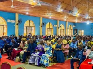 Large group of people seated in a bright blue hall with arched windows, attending a formal event; many wear colorful traditional clothing.