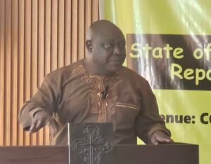 Man in a brown traditional outfit speaks at a podium, with a green banner in the background.