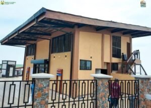 Two-story tan building with a wide overhanging roof behind a stone-pillared metal fence; a person stands near the gate with a staircase on the side.