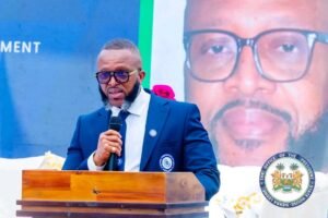 Man in a blue suit and glasses speaks into a microphone at a wooden podium during a formal event, with the speaker's portrait and an official seal in the background.