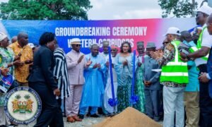 Officials and community members at a groundbreaking ceremony, with blue shovels and a sand mound, in front of a banner that reads 'Groundbreaking Ceremony' and the President's seal visible on the left.