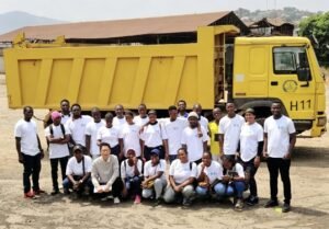 Group of people in white shirts posing together in front of a large yellow dump truck at a construction site or quarry.