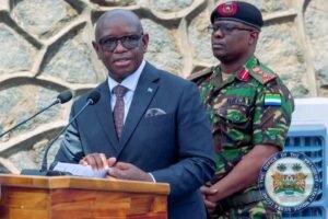 A man in a navy suit and patterned tie speaks at a wooden podium with two microphones; a military officer stands behind him.
