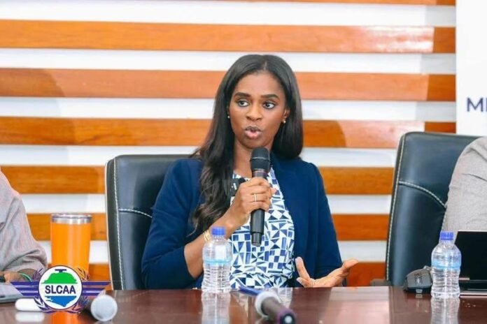 Professional woman in a blue blazer speaks into a handheld microphone during a panel discussion at a conference, seated at a table with water bottles and a logo in view.