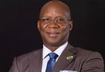Sierra Leone’s Mining Sector Hits USD 1.3 Billion in Exports, Signals Strong Economic Transformation Professional portrait of a smiling Black man in a brown suit, white shirt, and blue tie, against a dark background.