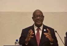 Health Minister Showcases Sierra Leone’s Progress on National Health Compact in Geneva Man in a dark suit and red tie speaks at a panel, gesturing with his hands; a microphone and water bottle are on the table beside him.