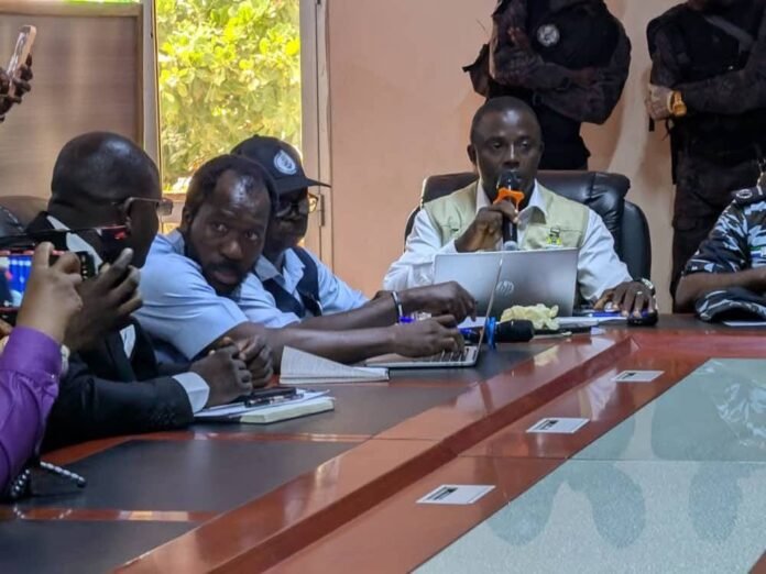 Man in a beige vest speaks into an orange microphone at a conference table, with colleagues on laptops nearby.