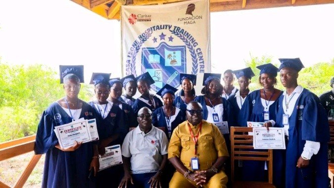 Group of graduates in blue gowns with certificates posing for a photo at a hospitality training center graduation event.