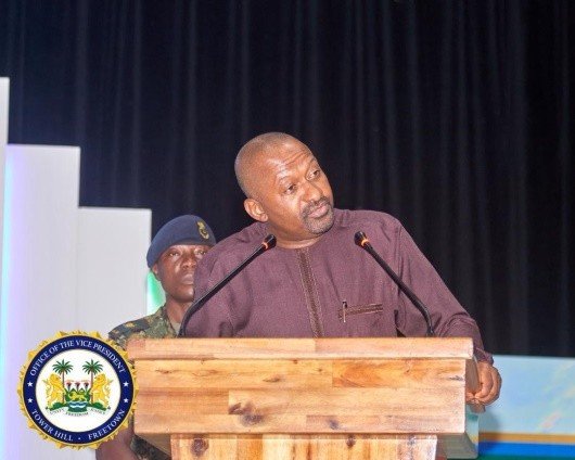Vice President, Dr. Mohamed Juldeh Jalloh Man speaks at a wooden podium on stage, with a uniformed guard behind and an official seal in the foreground left.