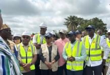 World Bank Endorses Manowa Bridge Progress, Highlights Impact on Farmers and Trade Group of construction workers and officials wearing hard hats and high-visibility vests listening to a briefing at a site.