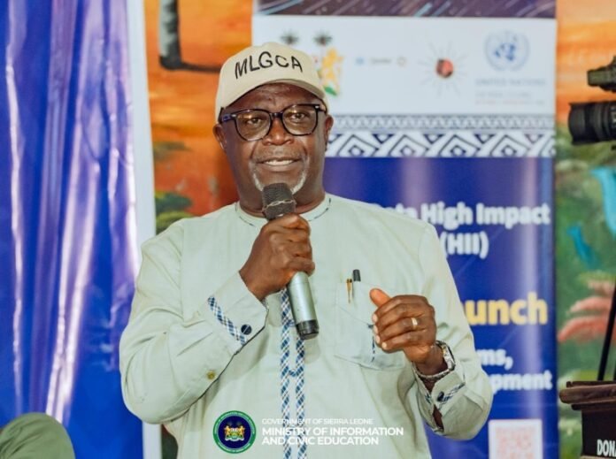 Man in a beige cap labeled 'MLGCA' speaks into a handheld microphone at a conference, banners in the background.