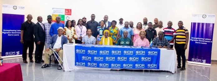 Group portrait of diverse officials and participants at an IOM event in Sierra Leone, seated at a table with IOM banners in front.