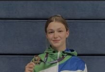 Josefien Betist Bags Third International Gold Medal in Six Months, Eyes Commonwealth Glory Young female athlete in a white tracksuit holding a gold medal, smiling against a blue wall.