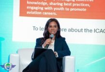 SLCAA DG Highlights Aviation Safety, Ebola Preparedness, Calls for Greater Public Cooperation in Lungi Woman in a dark blazer speaks into a handheld microphone while seated on a white chair, holding a folder, on a stage with a colorful presentation backdrop.