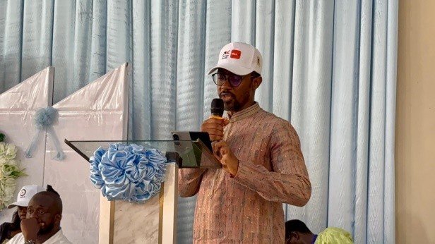 Man in a pink patterned shirt and white cap speaks into a microphone at a podium with a tablet, blue drapes behind him.