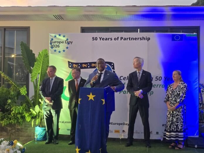 Five people in suits stand on a stage with a blue lectern decorated with EU stars at a Europe Day event titled '50 Years of Partnership.'