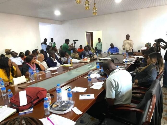 Diverse group seated around a long conference table for a meeting; journalists with cameras stand in the backroom area.