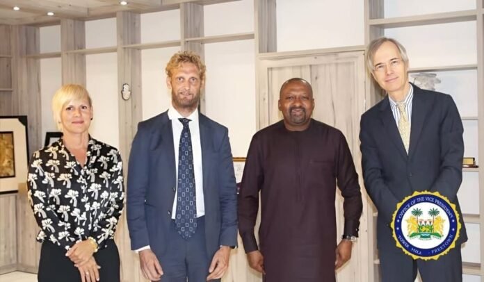 Four professionals pose for a photo in an office-like setting: a woman in a black floral blouse on the left, two men in suits, and a man in traditional dark attire, with an official seal for the Office of the Vice President visible at the bottom-right.