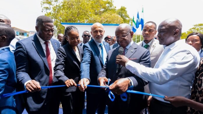 Officials in suits cut a blue ribbon at an outdoor ceremony, with onlookers and flags in the background.