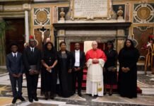 Ambassador Jusu Alongside Deputy Envoy Asmaa James Presents Credentials to Pope Leo XIV in Vatican City Eleven people in formal attire pose with a cardinal in red and white robes inside an ornately decorated church with marble floors and a crucifix backdrop.