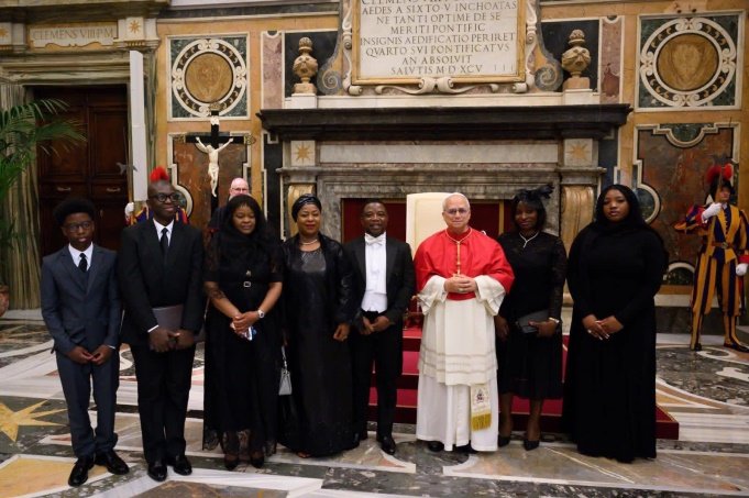 Eleven people in formal attire pose with a cardinal in red and white robes inside an ornately decorated church with marble floors and a crucifix backdrop.