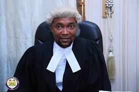 Judge seated in a courtroom, wearing a black robe with white neck bands and a serious expression.